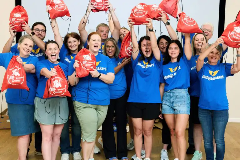 Joyful group of adults in blue 'changemakers' shirts raise red drawstring bags in celebration indoors.